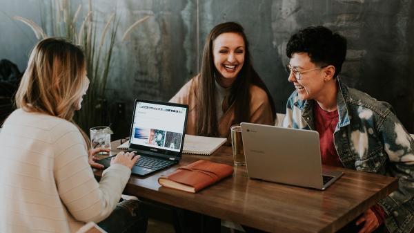 Three people sit around a wooden table, all with laptops laughing.