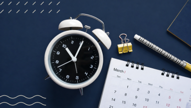 An old fashioned alarm clock is beside a calendar on a dark blue table.